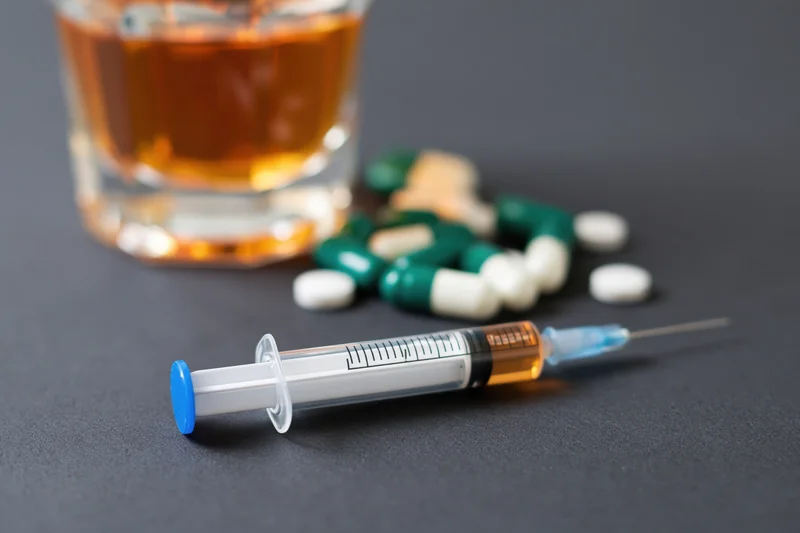Syringe with blue cap lying on dark matte surface beside glass of amber liquid and scattered pills