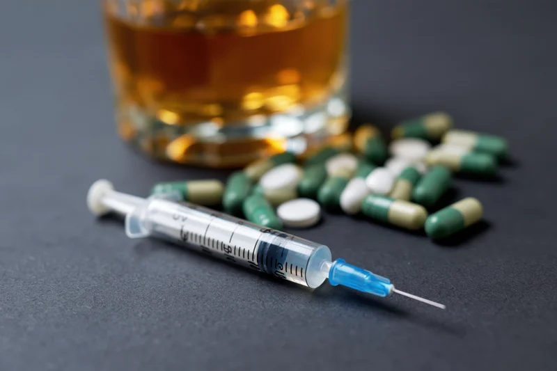 Syringe with blue cap lying on dark surface beside glass of amber liquid and scattered pills