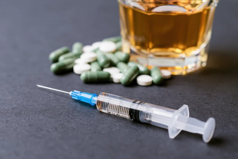 Syringe with blue cap lying on dark surface beside glass of whiskey and scattered pills