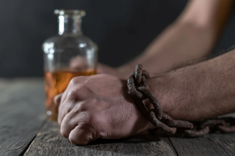 A man’s hand bound by a rusted metal chain resting on a wooden table beside a glass bottle of whiskey
