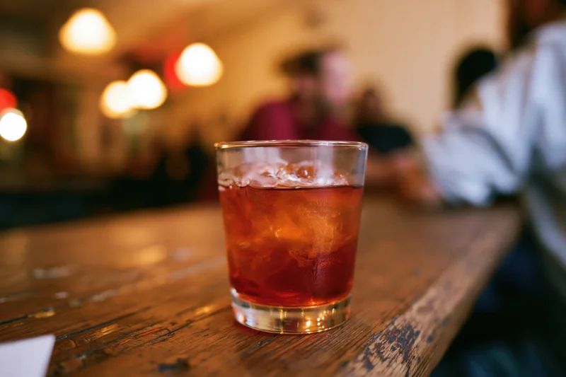 Amber-red cocktail with ice in a rocks glass on a wooden bar table