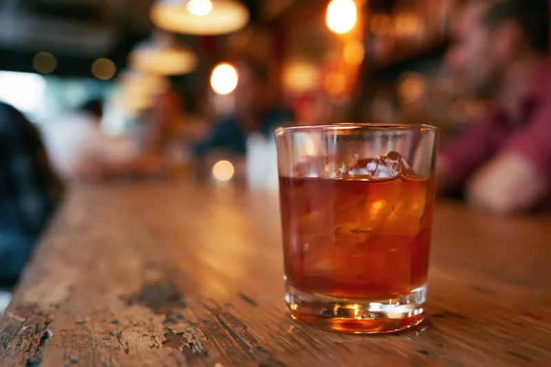 Amber-red cocktail with ice in a rocks glass on a rustic wooden bar table