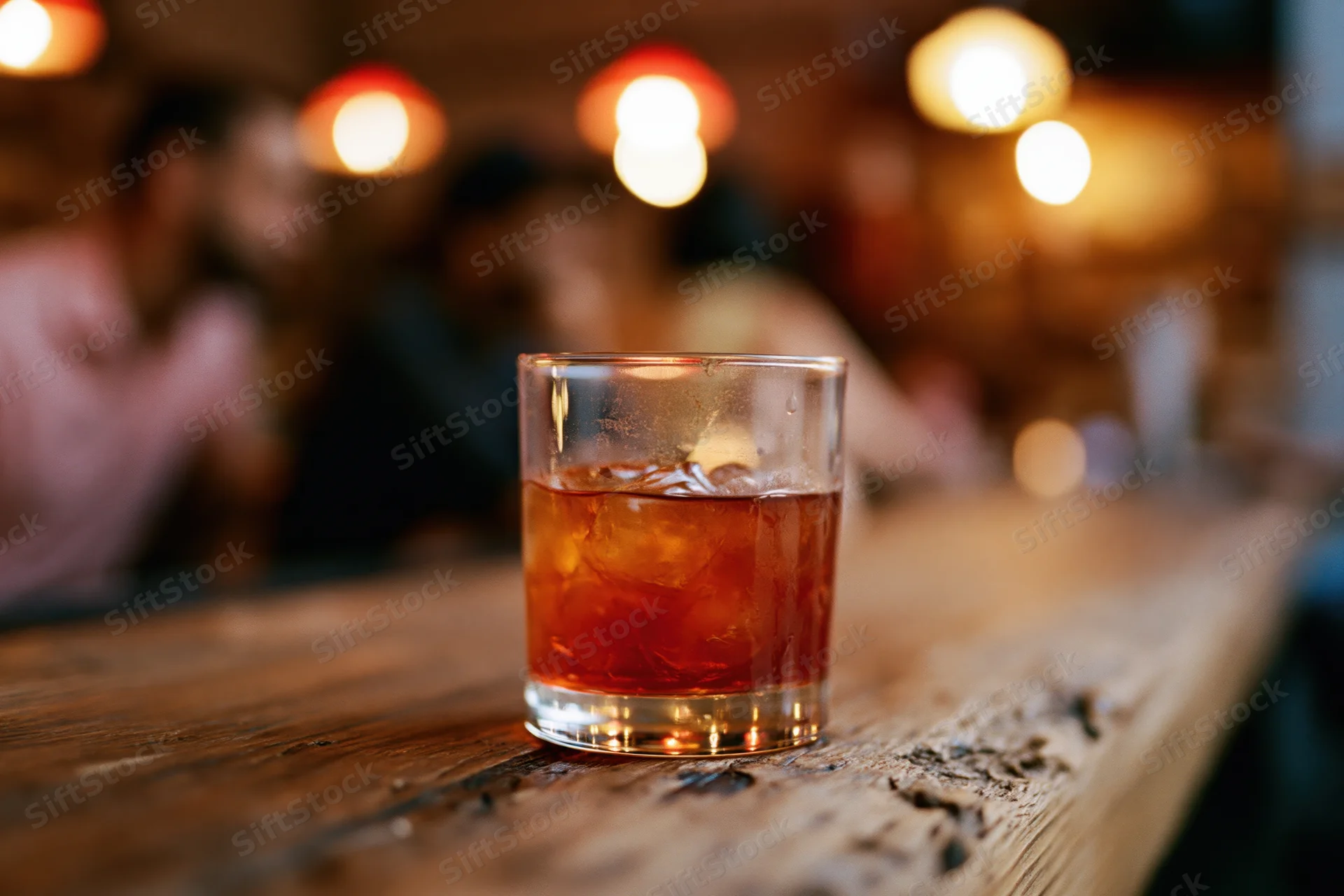 Amber-red cocktail with ice in a rocks glass on a wooden bar countertop, dimly lit bar background with blurred people and warm bokeh lights