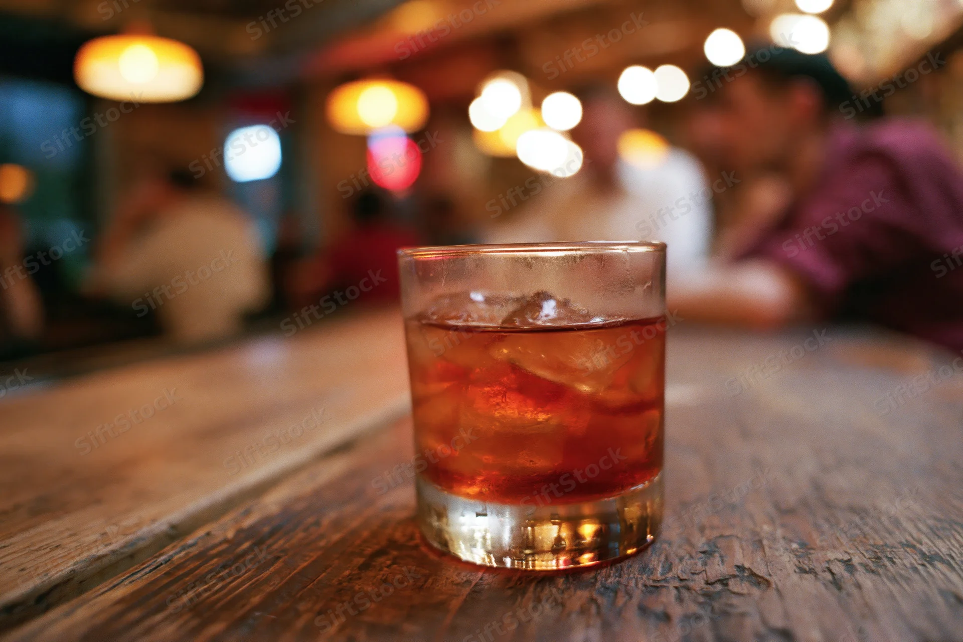 Amber-red cocktail with ice in a rocks glass on a wooden bar table in a dimly lit bar