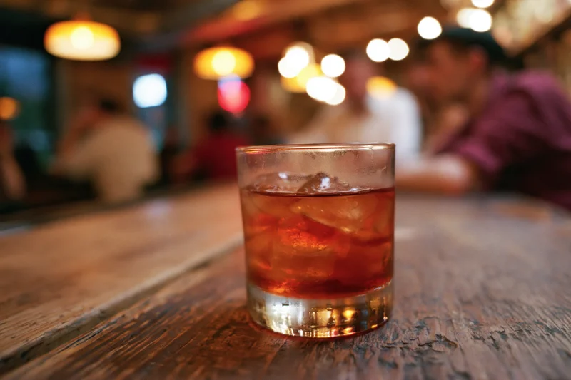 Amber-red cocktail with ice in a rocks glass on a wooden bar table in a dimly lit bar