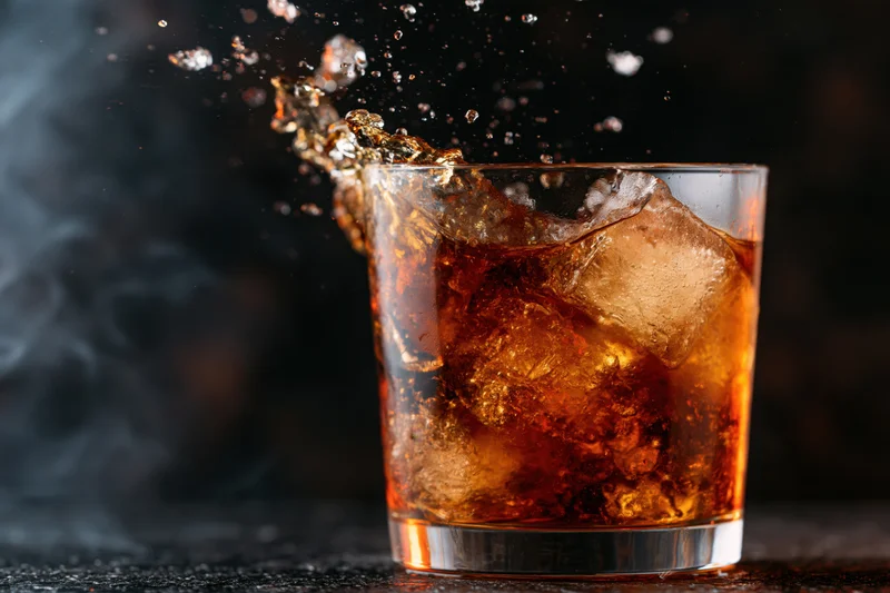 Dark amber whiskey in a rocks glass with ice cubes splashing against a black background