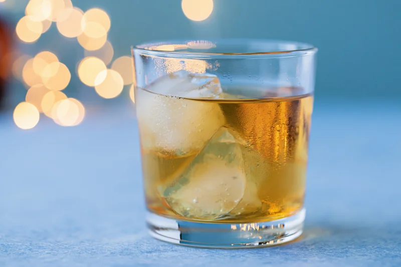Whiskey in a rocks glass with ice on a blue surface, bokeh lights in background