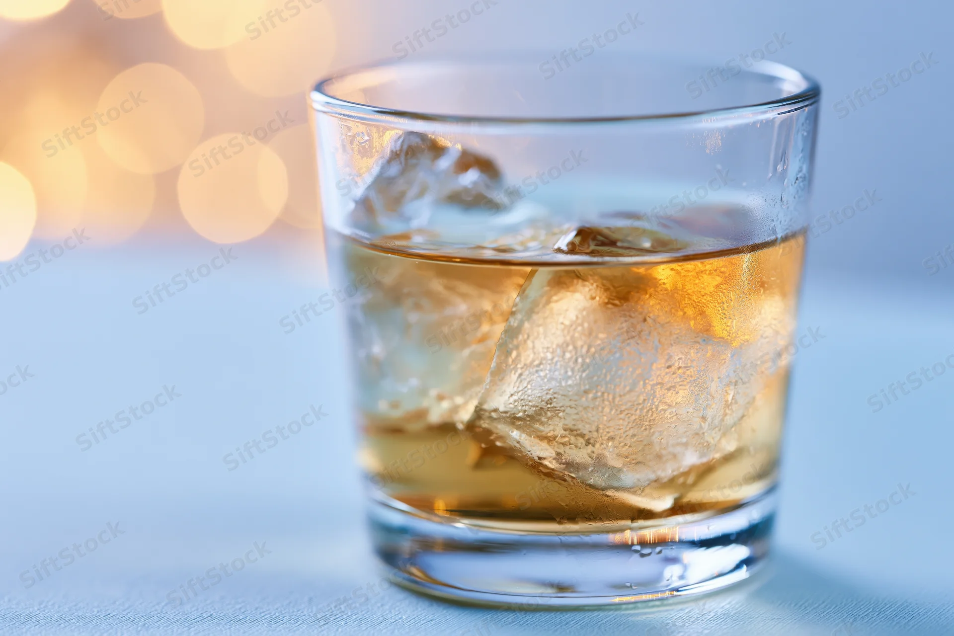 Whiskey in a rocks glass with ice on a blue surface against warm bokeh lights