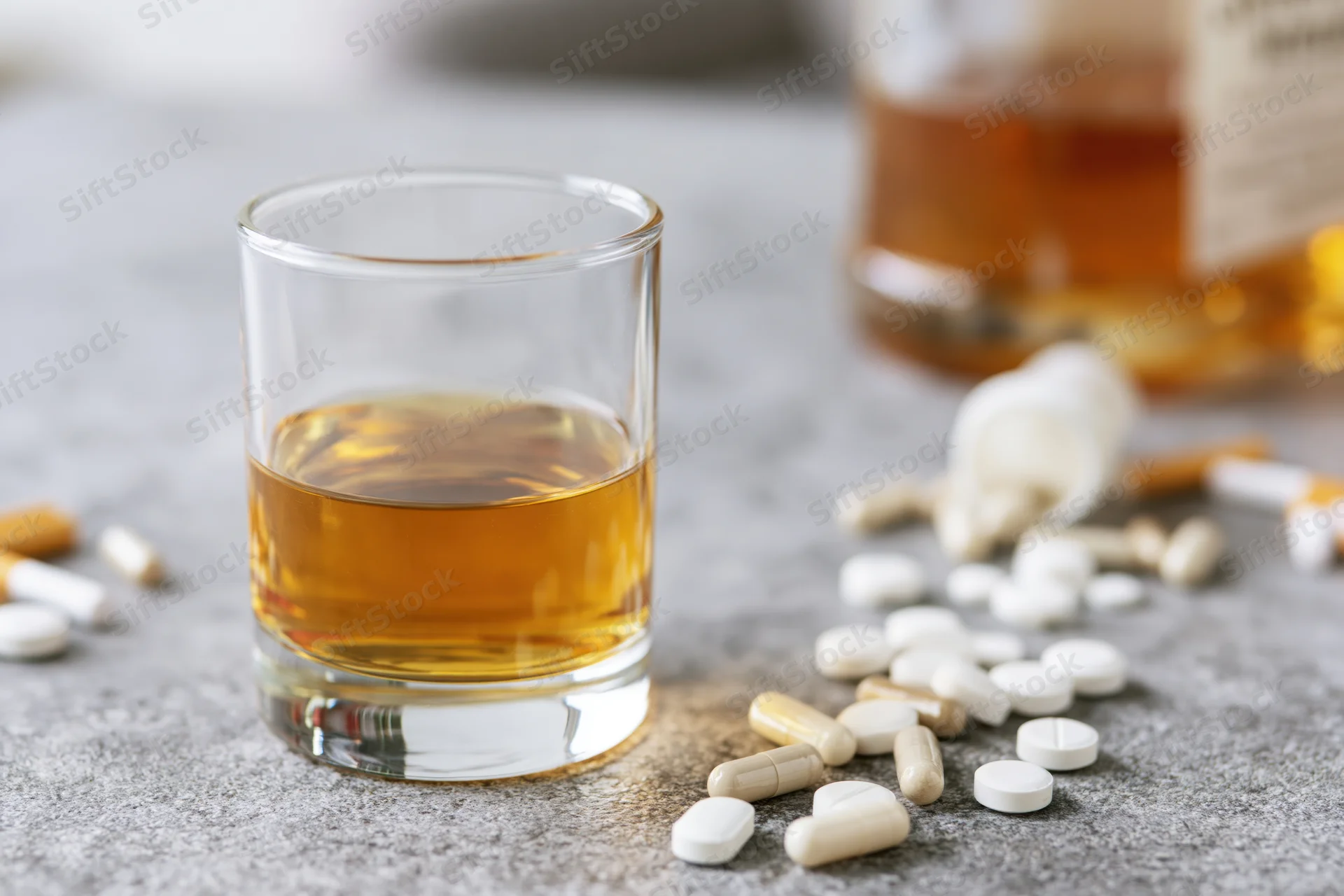 Half-filled whiskey glass with scattered pills on a grey countertop