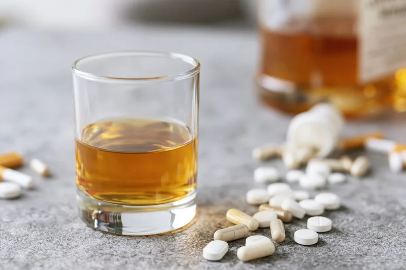 Half-filled whiskey glass with scattered pills on a grey countertop