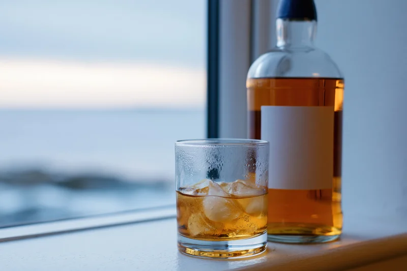 Whiskey in a rocks glass with ice beside a bottle on a windowsill overlooking the ocean at sunset