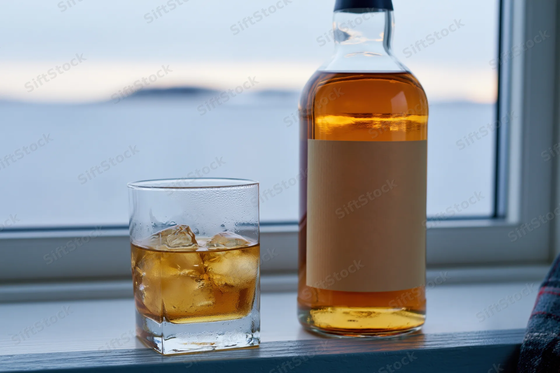 Whiskey bottle and rocks glass with ice on a windowsill by the sea at twilight