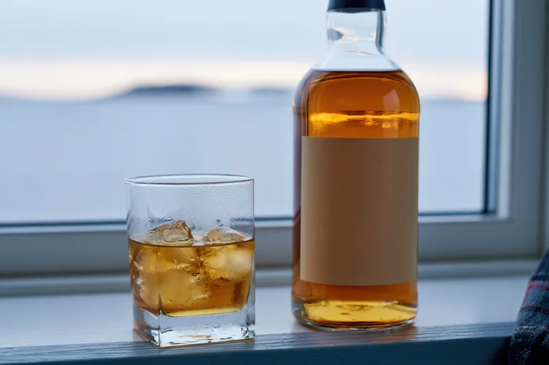 Whiskey bottle and rocks glass with ice on a windowsill by the sea at twilight