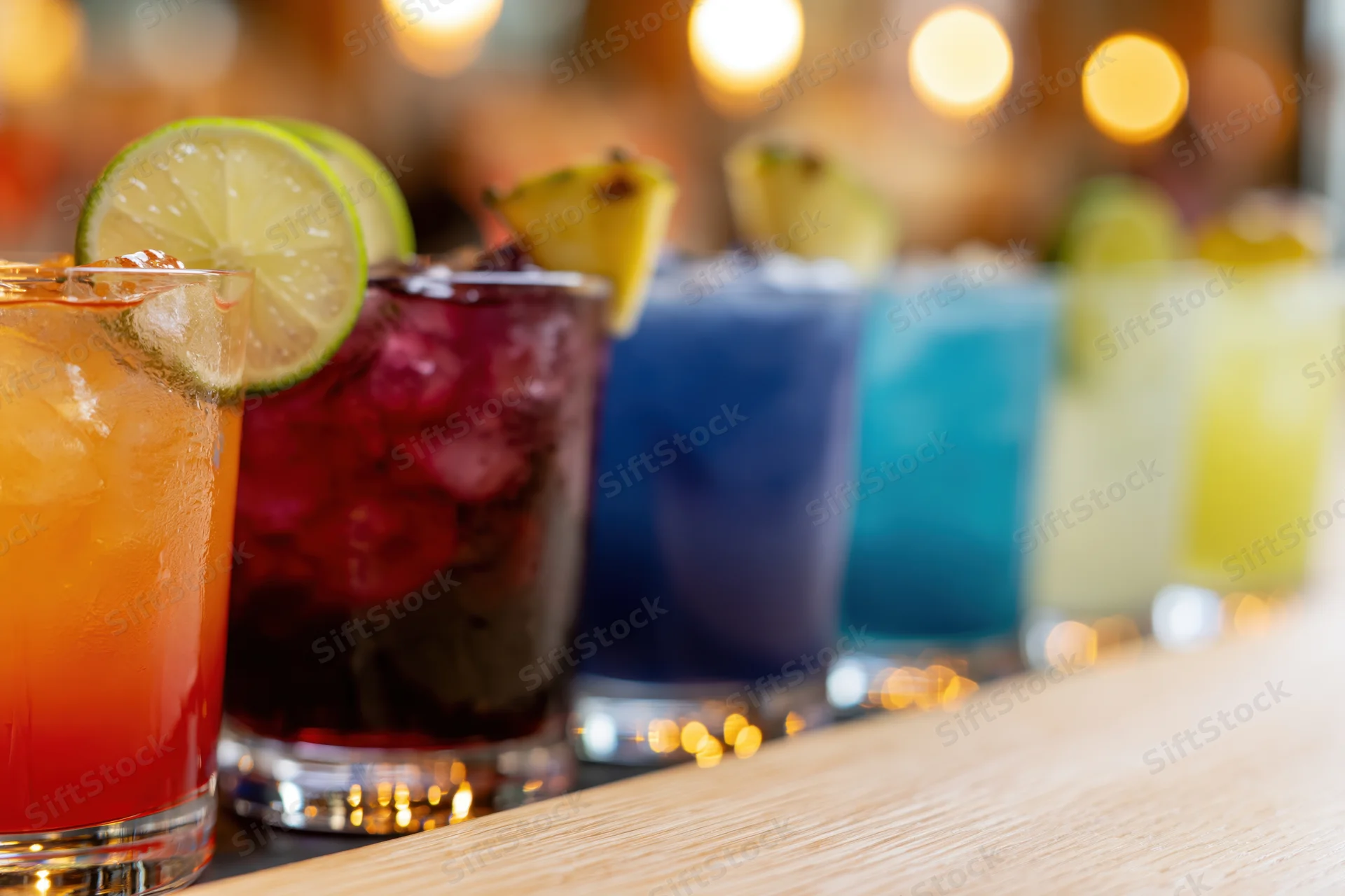 Row of colorful cocktails with ice and citrus garnishes lined up on a wooden bar counter