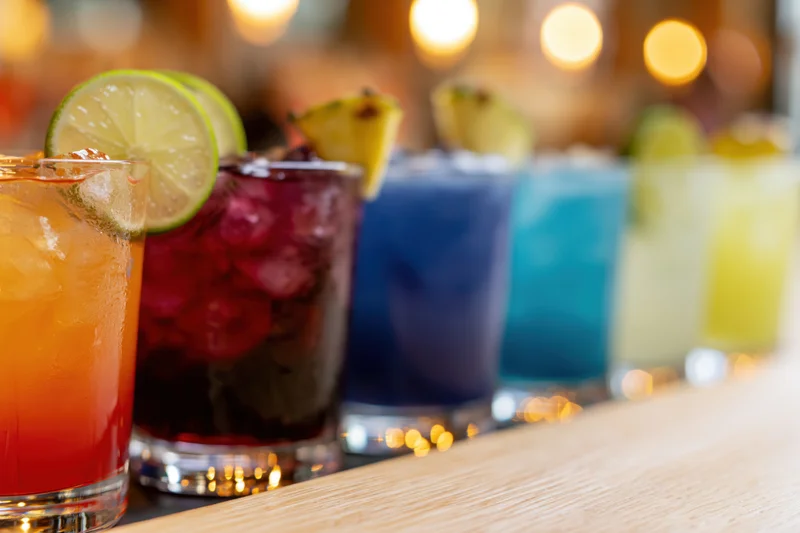 Row of colorful cocktails with ice and citrus garnishes lined up on a wooden bar counter