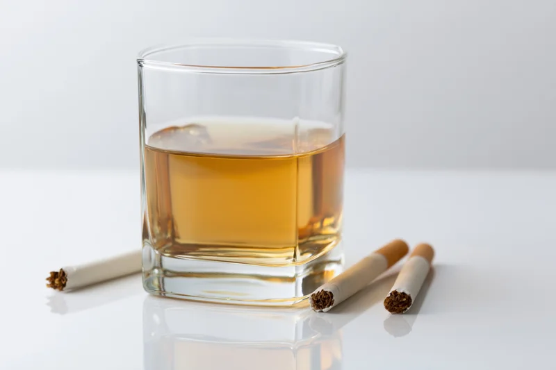 A square glass of amber bourbon with three cigarettes on a white reflective surface
