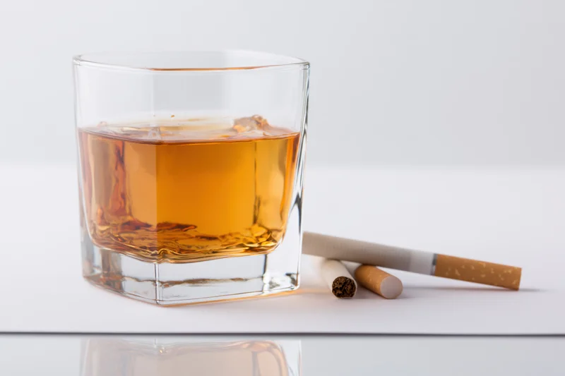 Square whiskey glass with amber bourbon and cigarettes on white surface