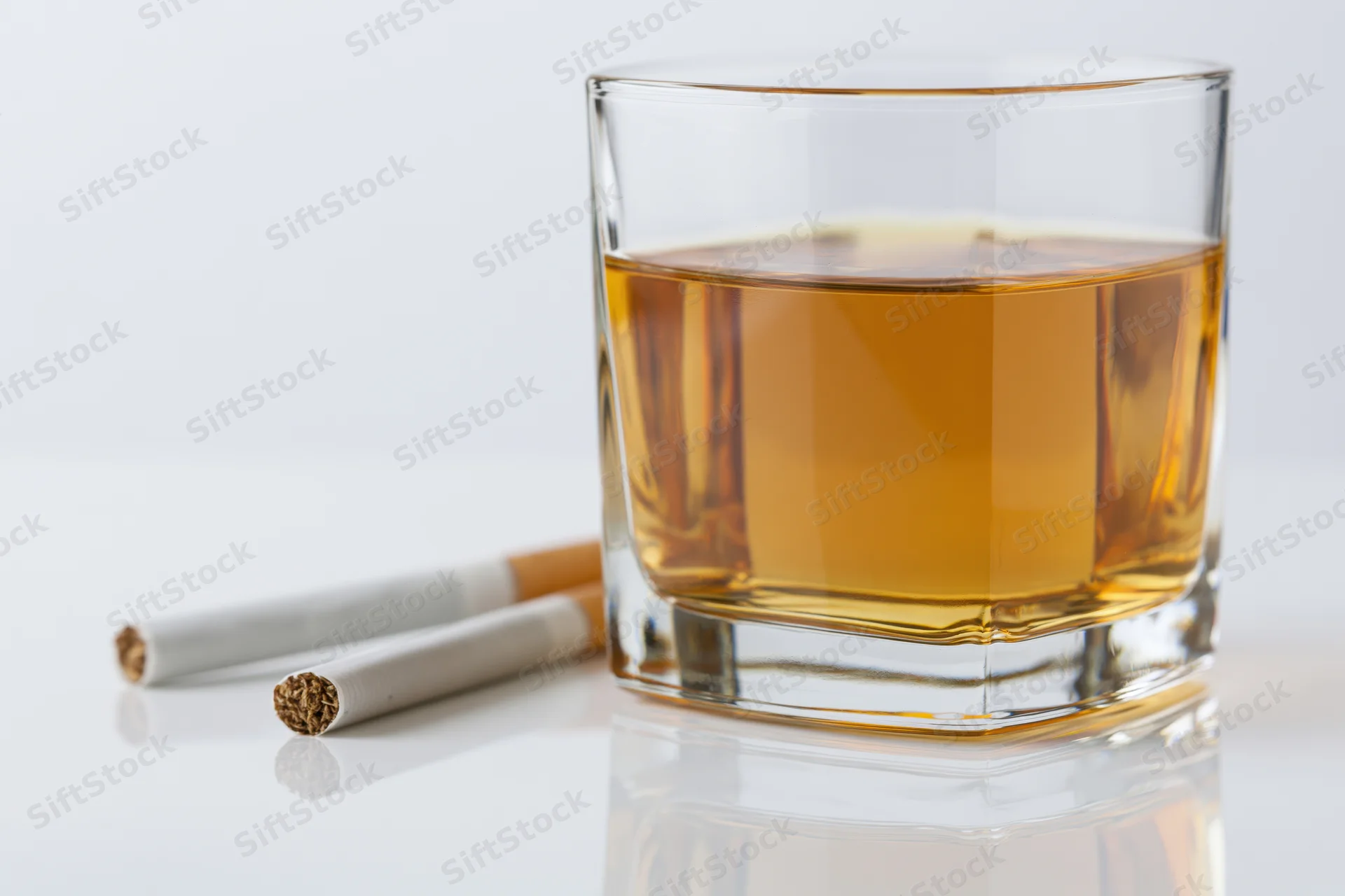 Whiskey glass filled with amber bourbon beside two cigarettes on a white reflective surface