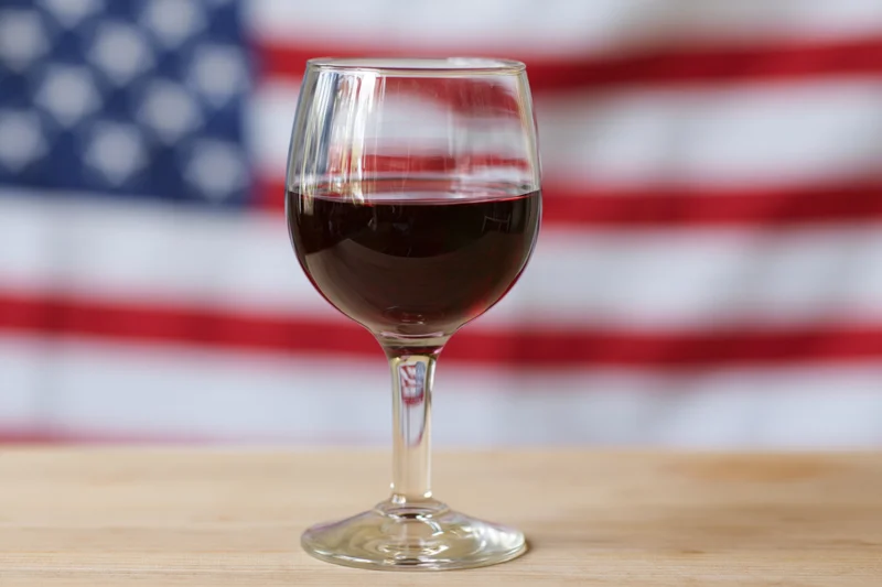 A wine glass filled with red wine on a wooden table with american flag in background