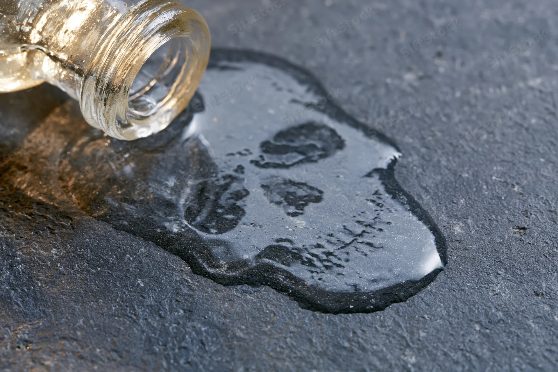Glass bottle tipped over spilling liquid onto dark pavement surface