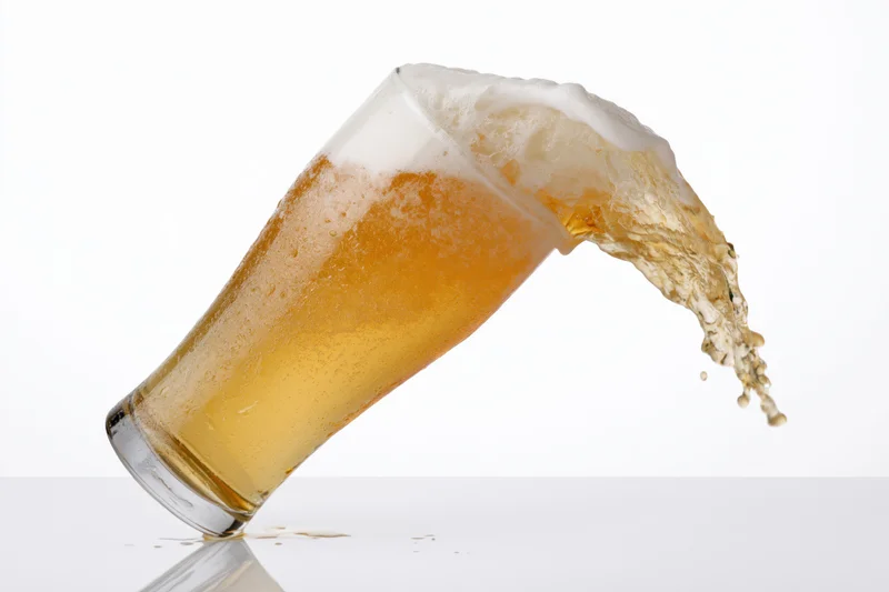 Tilting glass of lager splashing beer liquid on white surface