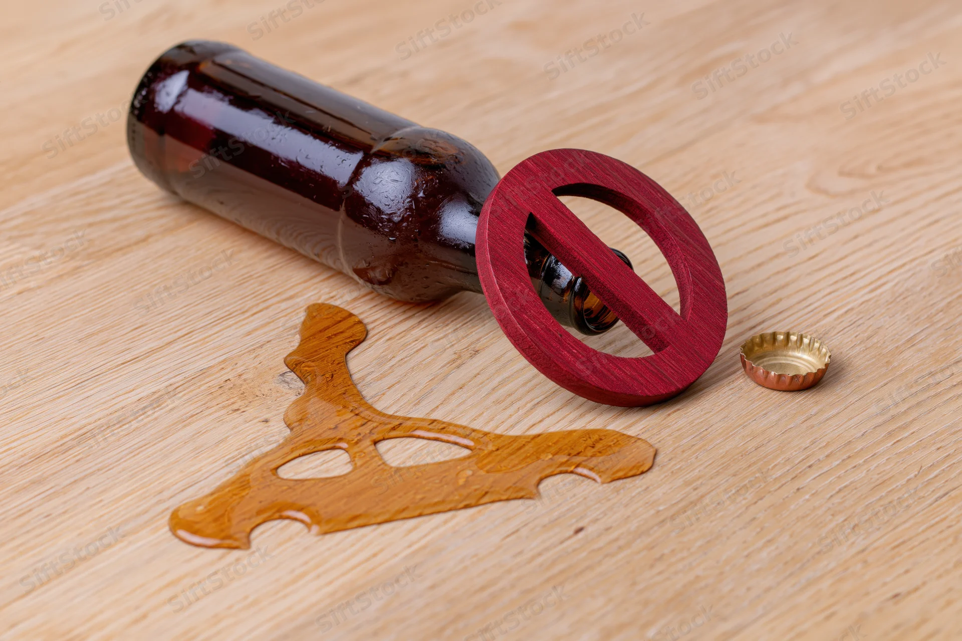 Tipped beer bottle with spilled liquid and red prohibition symbol on wooden table