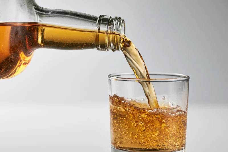 Amber whiskey being poured from a tilted glass bottle into a clear glass tumbler on a white background