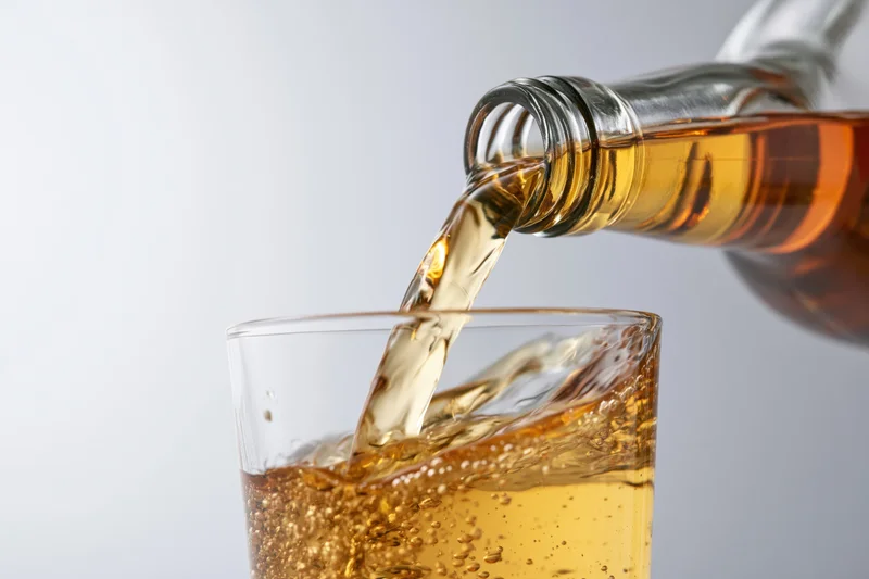 Amber whiskey being poured from a tilted glass bottle into a tumbler glass against a plain background