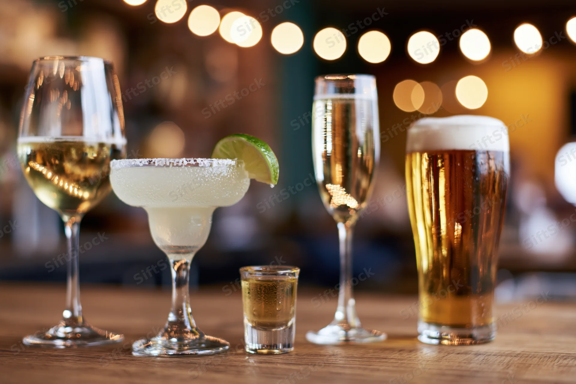 Assorted alcoholic drinks lined up on a wooden bar counter with string lights in the background