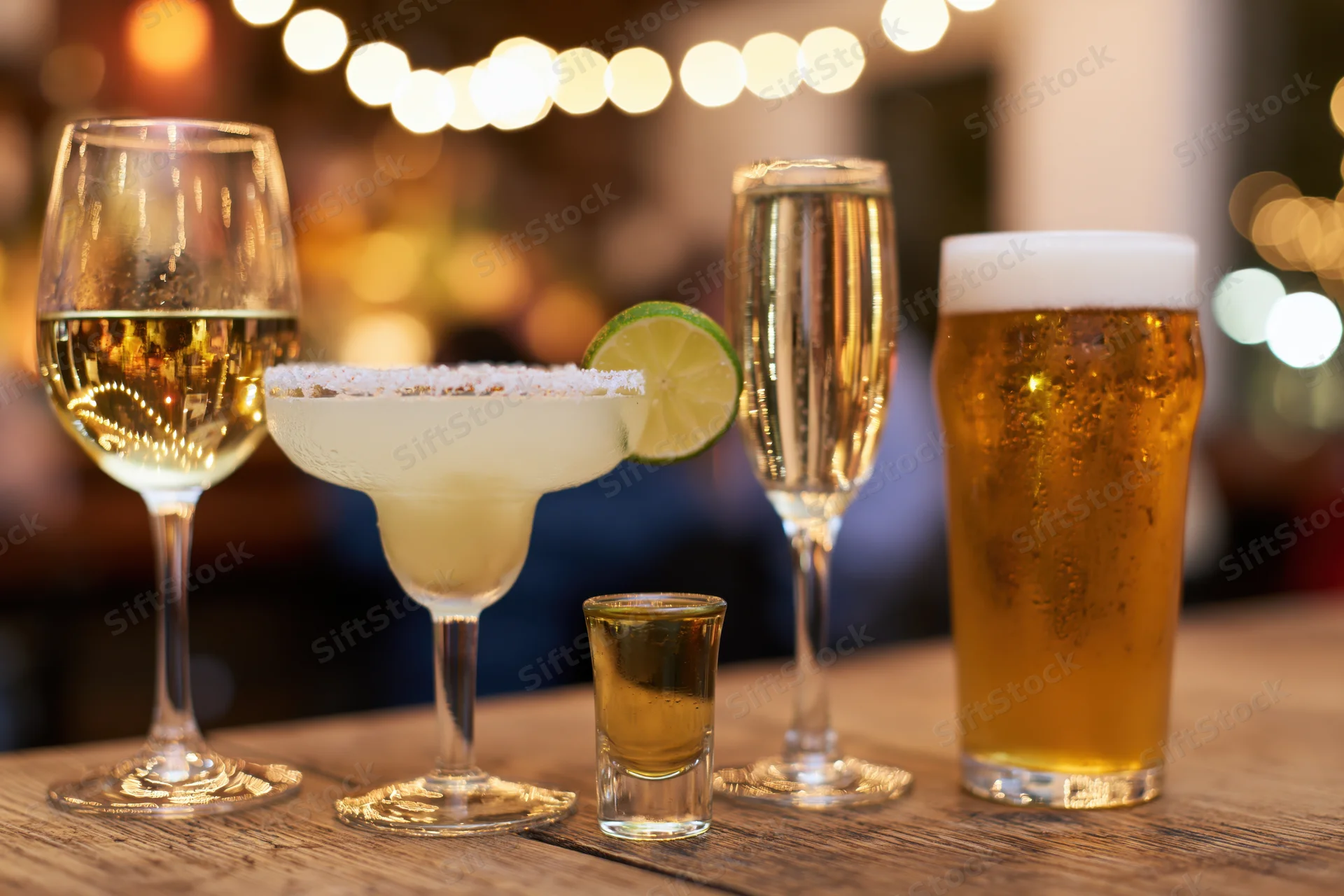 Assorted alcoholic drinks lined up on a wooden bar counter with string lights in the background