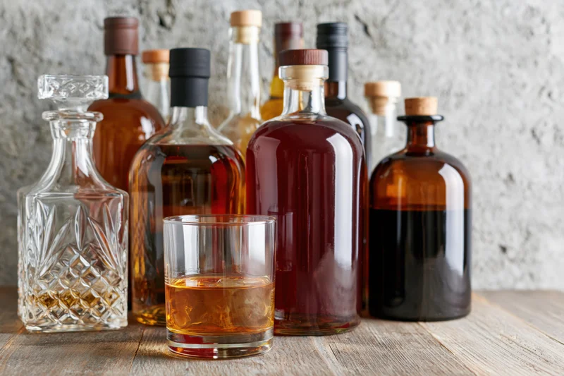 Assorted liquor bottles and a crystal decanter arranged on a wooden table with a glass of whiskey and ice