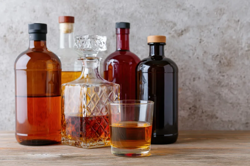 Assorted liquor bottles and a crystal decanter arranged on a wooden table with a glass of amber liquid in front