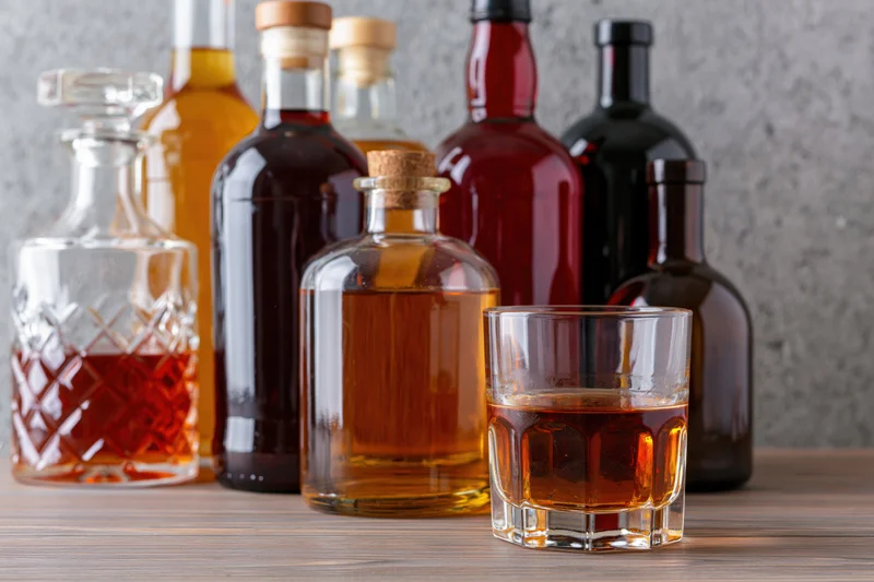 Whiskey in a tumbler with assorted liquor bottles and a crystal decanter on a wooden table