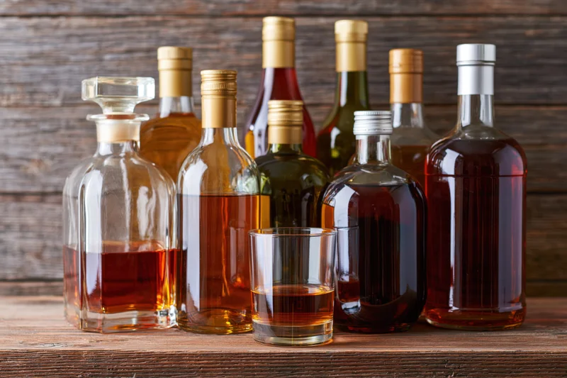 Assorted liquor bottles and a glass decanter arranged on a wooden tabletop with a tumbler filled with amber liquid in front