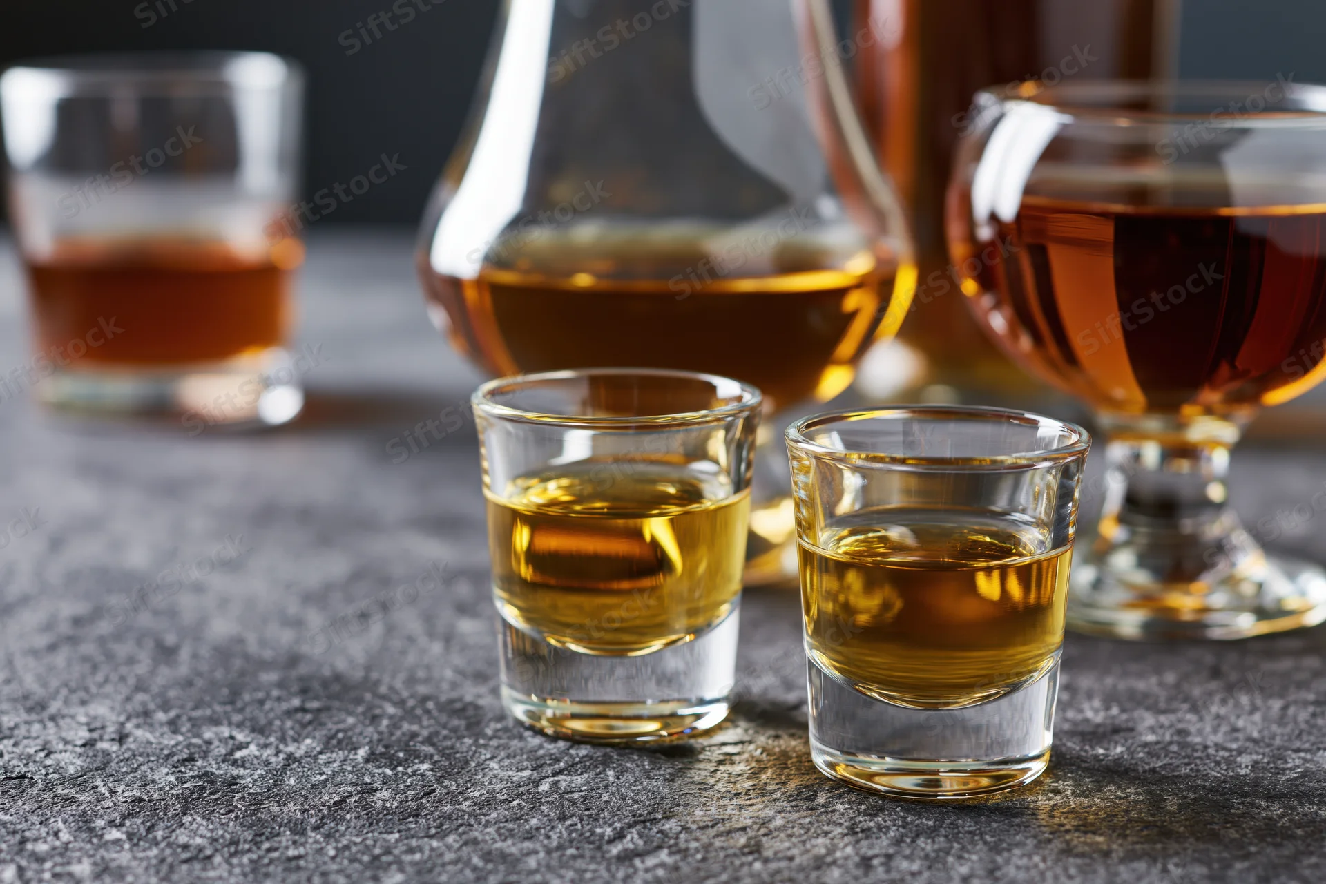 Whiskey and cognac glasses arranged on a dark wet countertop with decanter and shot glasses in focus