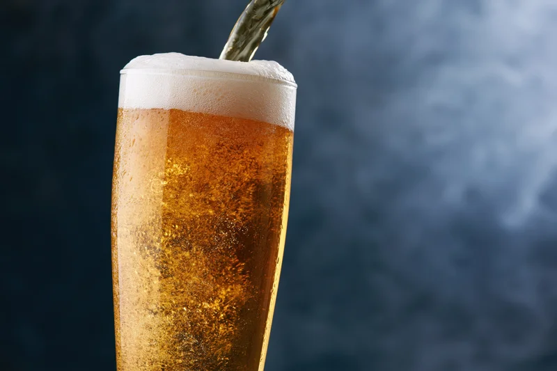 Golden lager being poured into a tall pilsner glass with foam and condensation against a dark background