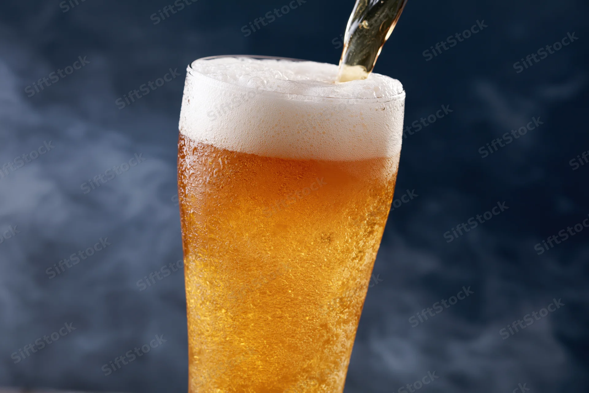 Golden lager being poured into a tall pilsner glass with foam forming on top against a dark background