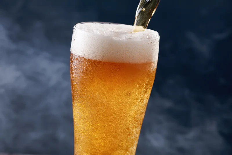 Golden lager being poured into a tall pilsner glass with foam forming on top against a dark background