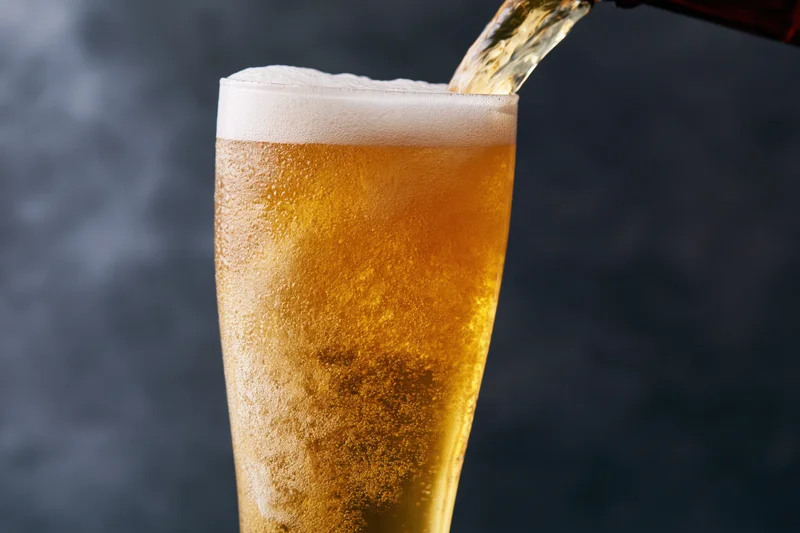 Golden lager being poured into a tall pilsner glass with foam forming on top against a dark background