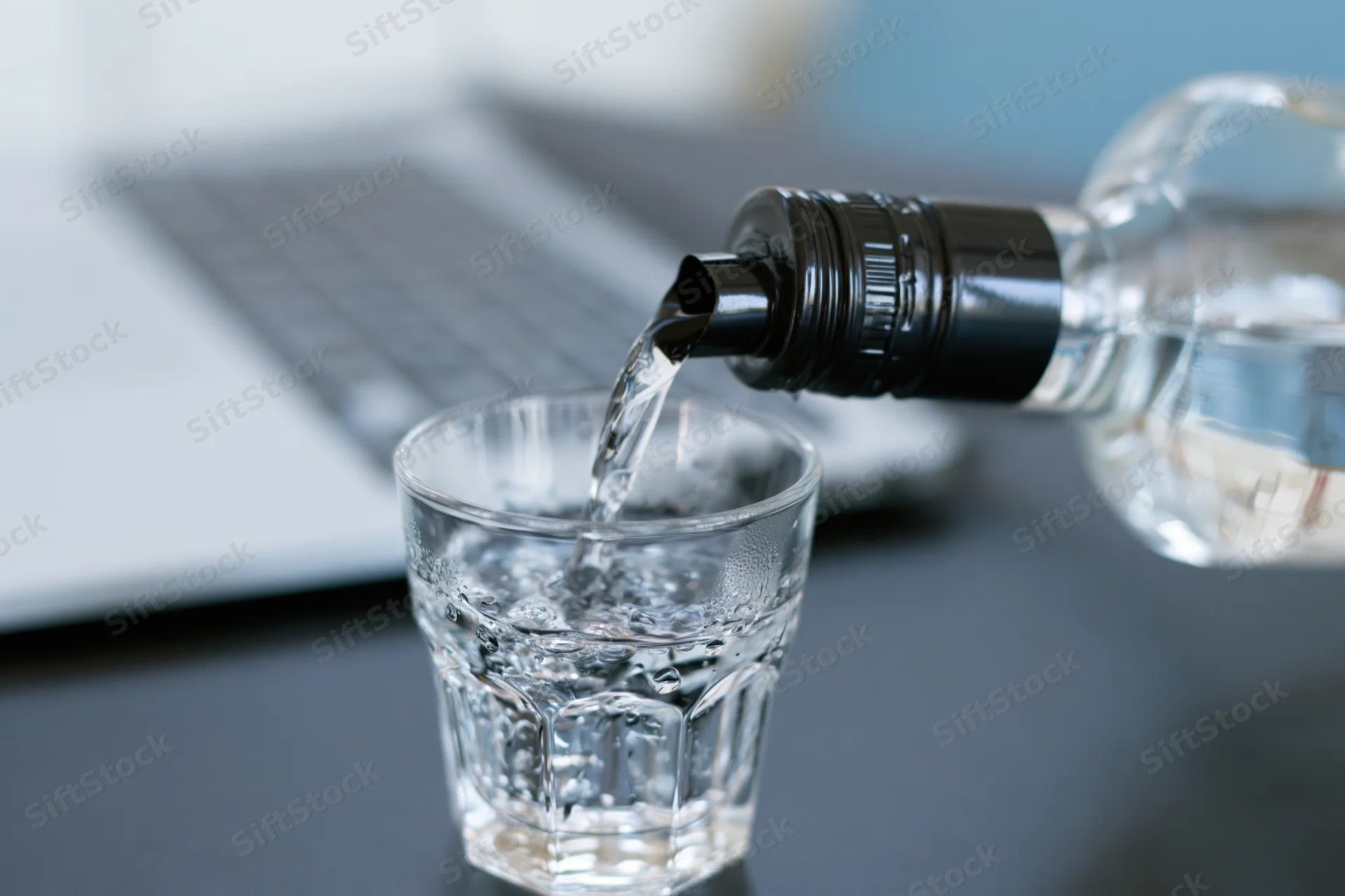 Water being poured from a bottle with a black spout into a glass on a desk near a laptop