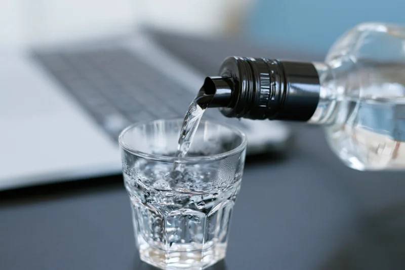 Water being poured from a bottle with a black spout into a glass on a desk near a laptop