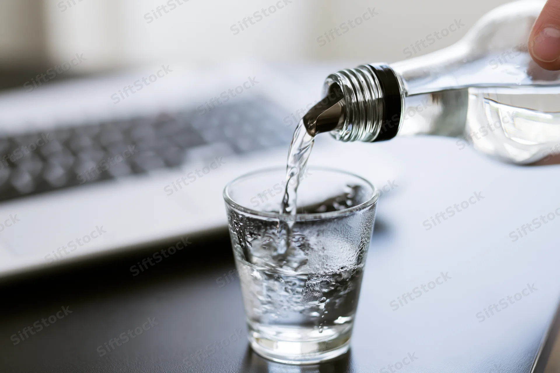 Clear glass bottle pouring vodka into a shot glass on a desk beside a laptop