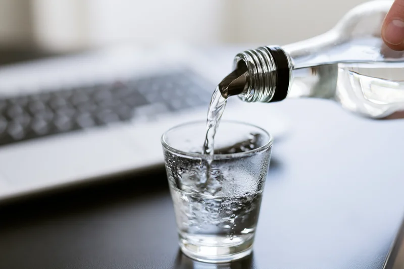 Clear glass bottle pouring vodka into a shot glass on a desk beside a laptop