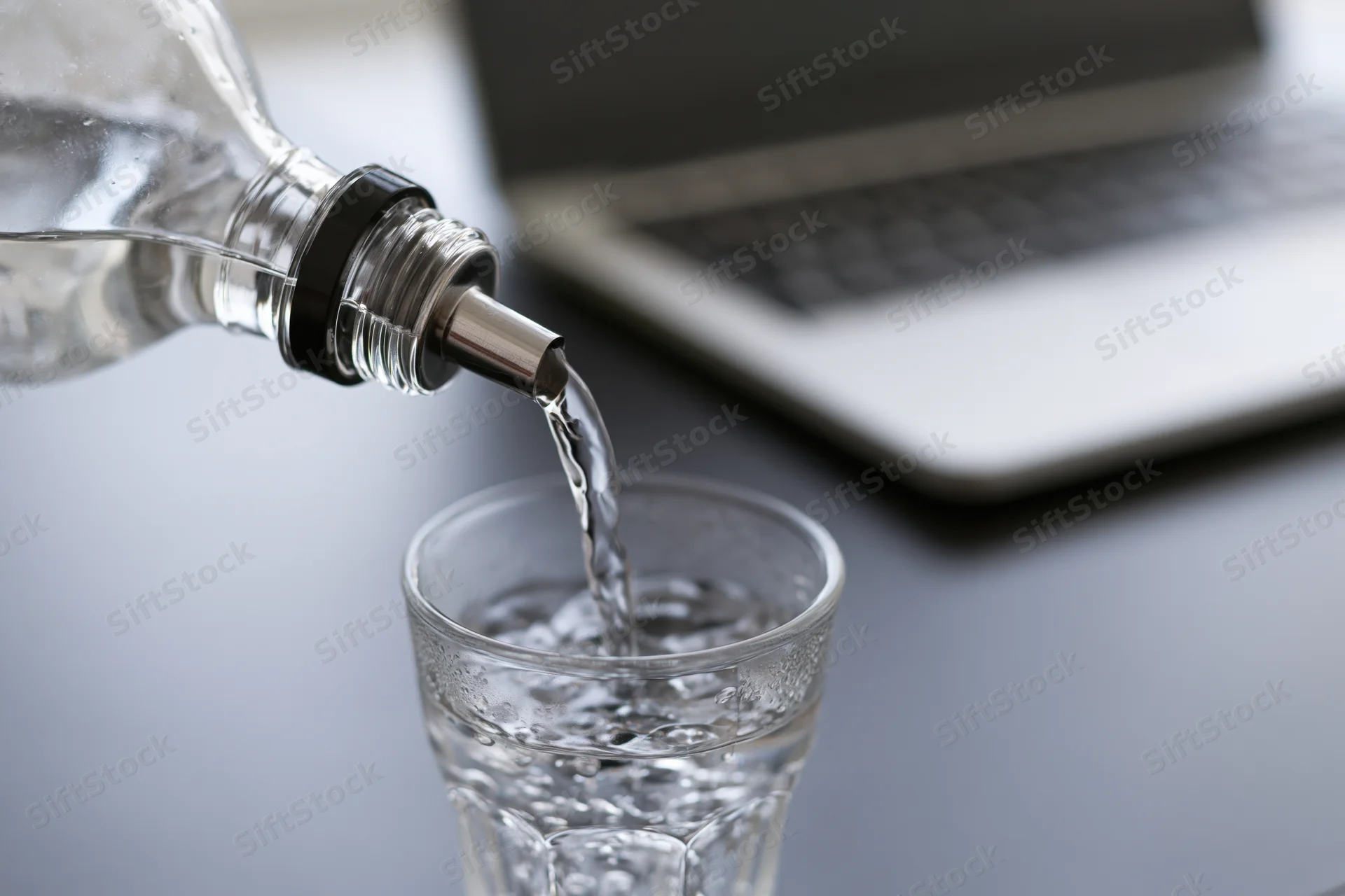 Water being poured from a clear glass bottle into a glass on a desk beside a laptop