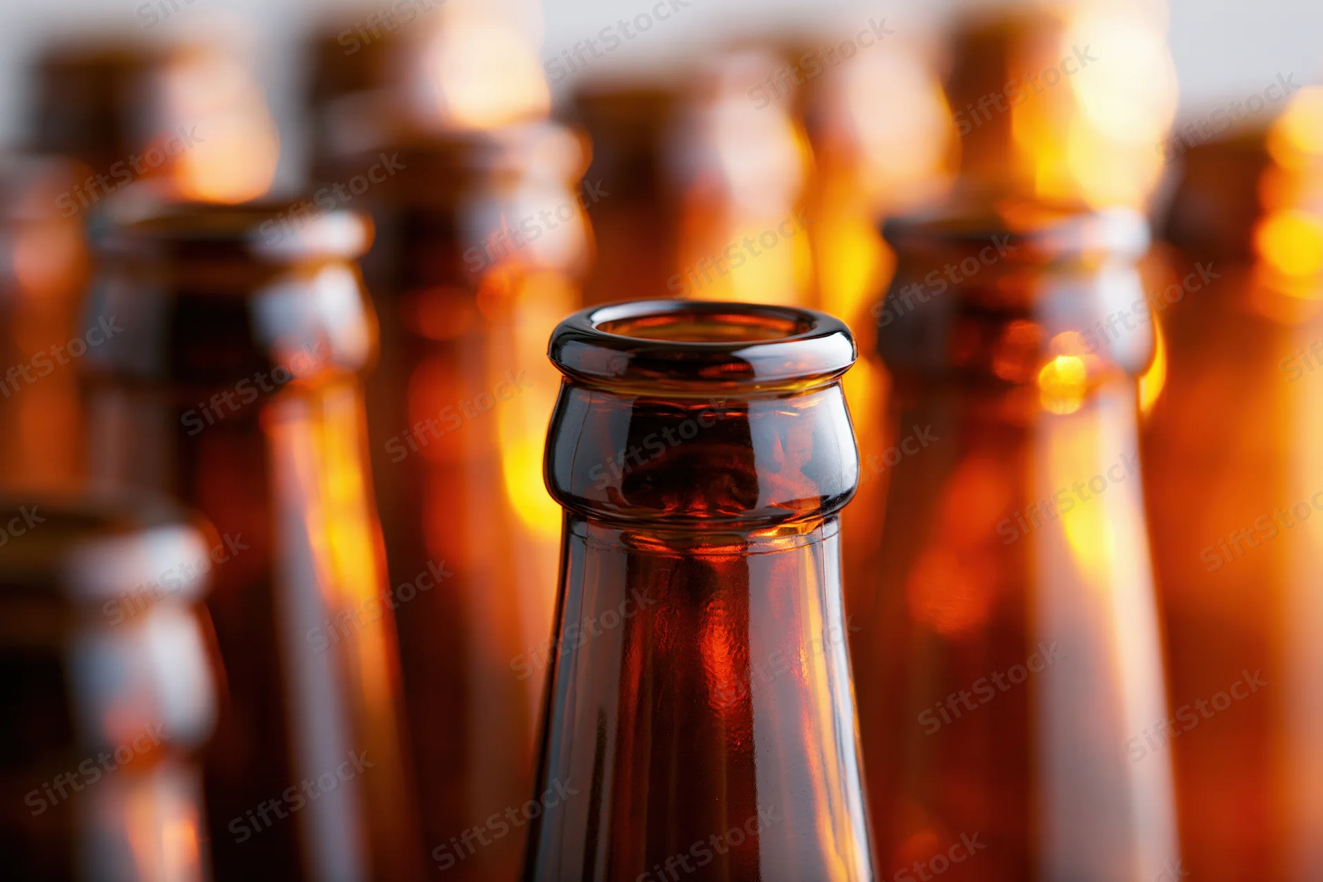 Close-up of empty amber glass beer bottle necks arranged in rows with warm light reflections