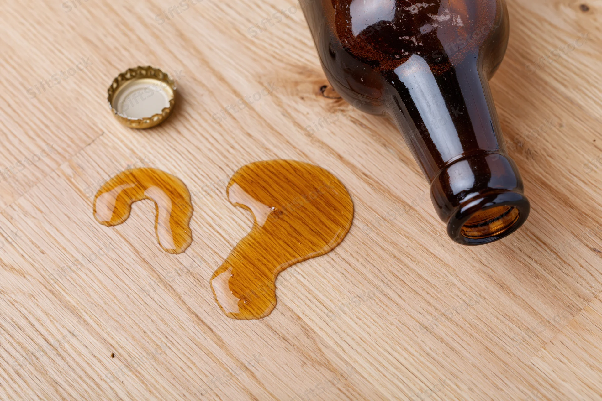 Dark brown glass beer bottle lying on its side on light wooden surface with spilled liquid forming question mark shape and detached cap nearby