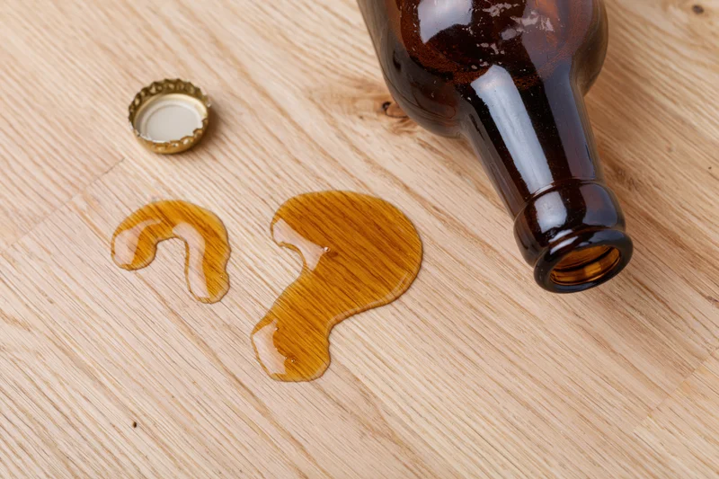 Dark brown glass beer bottle lying on its side on light wooden surface with spilled liquid forming question mark shape and detached cap nearby
