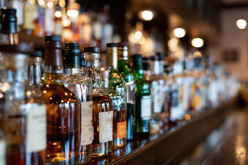 Assorted liquor bottles lined up along a dark wooden bar counter with warm ambient lighting and bokeh background