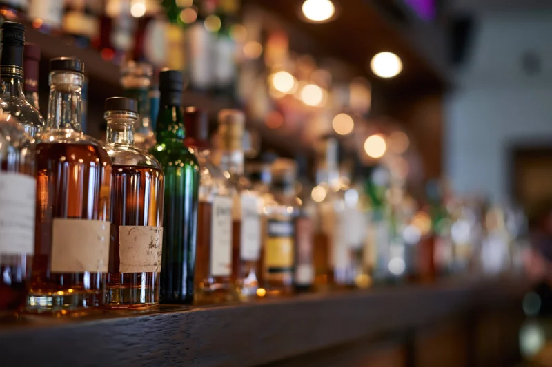 Assorted liquor bottles lined up on a wooden bar shelf in a dimly lit bar interior