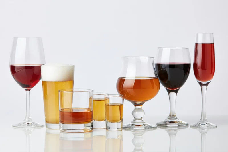 Assorted drink glasses filled with different alcoholic beverages arranged in a row on a reflective white surface